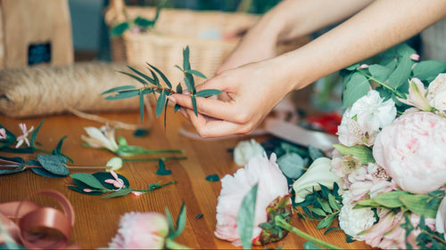 A close-up lifestyle shot of a florist's hands arranging fresh green leaves and stems on a wooden table surrounded by pink and white peonies, roses, and floral supplies.