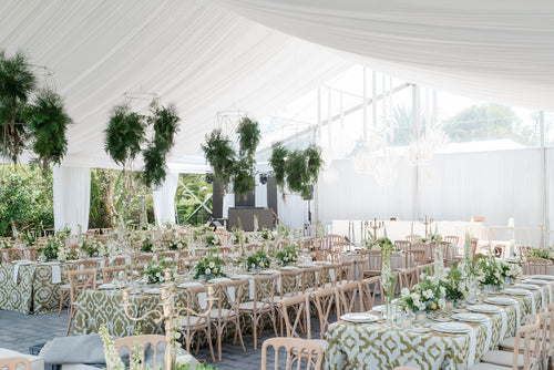 A large white event tent set up for a reception or wedding reception, with multiple tables covered in green and white patterned tablecloths, white floral centerpieces, and light wood chairs. Tall hanging greenery decorates the white draped ceiling.