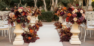 Wedding aisle with sall vessels and lower arrangements in fall color flowers(garden roses, standard rose, dahlias, hydrangeas, silver dollar eucalyptus and more), brown, burgundy, peach, orange and red. White chairs and a white floor runner. Bloomingmore