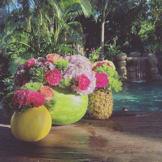 DIY summertime fruit vases made from hollowed watermelon honeydew and pineapple filled with fresh carnations and hydrangeas as creative party centerpieces