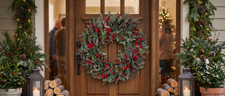 A festive Christmas wreath with red berries and green foliage hangs on a wooden front door, flanked by decorated garlands, lanterns, and stacked logs in a warmly lit entryway.